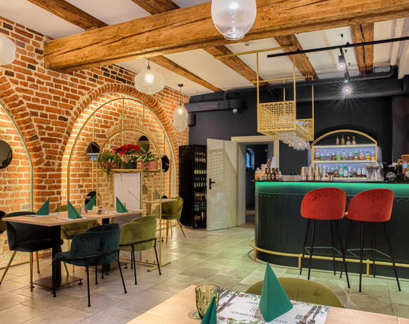 Nowoczesny bar hotelowy z ekspozycją alkoholi, ciepłym oświetleniem, czerwonymi hokerami i przytulnymi miejscami przy stole.