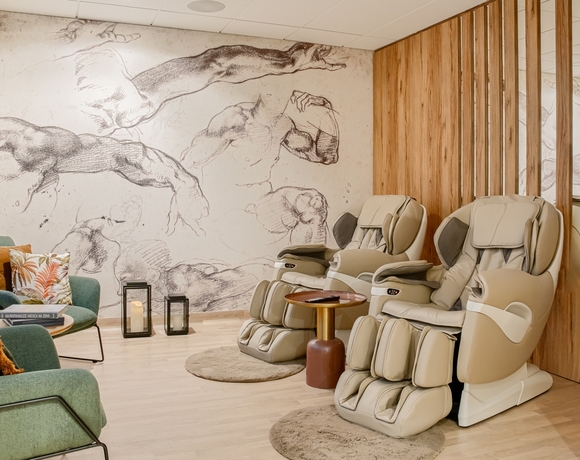 Relaksujący pokój spa z dwoma fotelami do masażu, zielonymi fotelami i artystyczną ścianą z rysunkami ciał ludzkich.