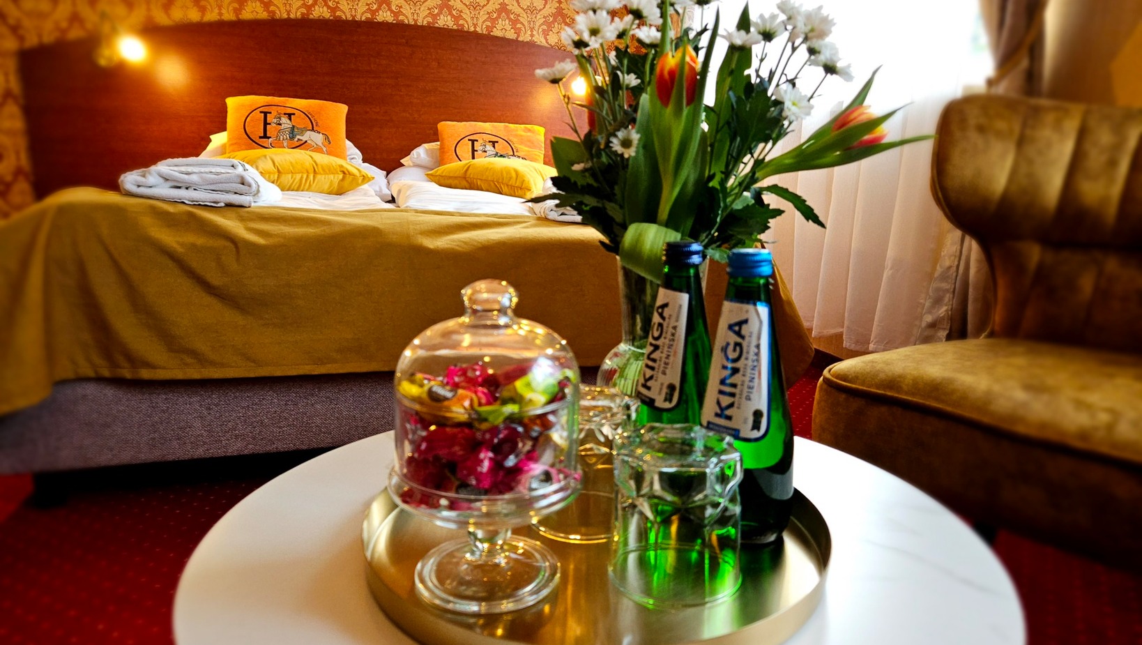 Elegancki pokój hotelowy z podwójnym łóżkiem, złotą narzutą, kwiatami i butelkami wody mineralnej na stoliku