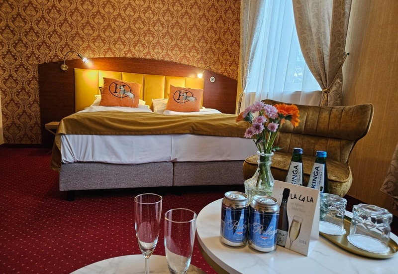 Letto matrimoniale con testiera gialla imbottita, carta da parati classica, lampade da lettura, poltrona, fiori freschi e acqua minerale.