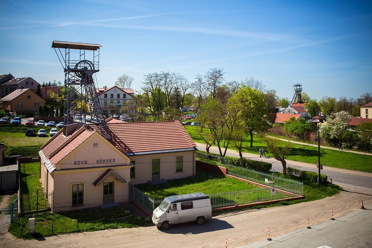 Stara wieża szybowa i budynek z czerwoną dachówką przy parku, zielone drzewa i zaparkowany van na tle niebieskiego nieba