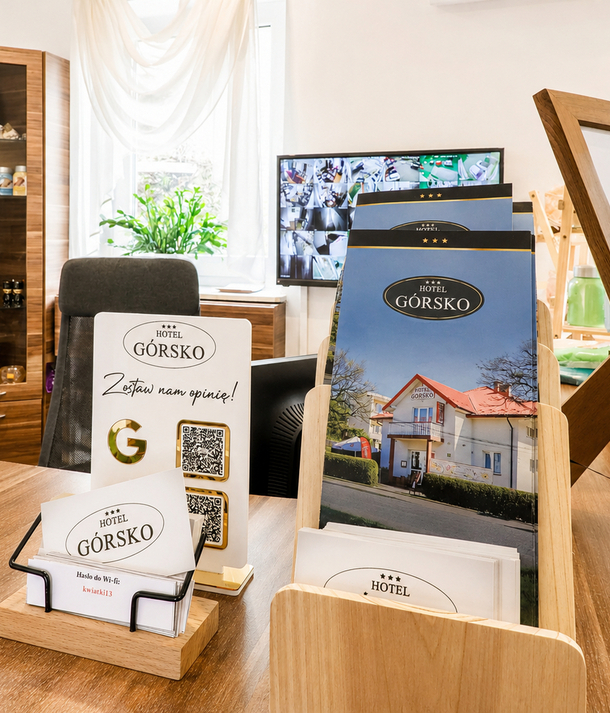 Recepcja Hotelu Górsko: drewniany blat z broszurami, wizytówkami z hasłem Wi‑Fi, tablicą meldunkową i monitorem CCTV.
