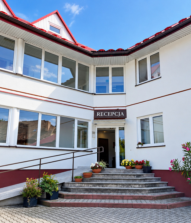 Wejście do recepcji hotelu z podjazdem i schodami, donice z kwiatami, biały budynek z czerwonym dachem
