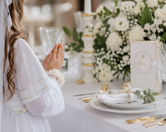 Dziewczynka w białej sukni komunijnej modli się przy elegancko nakrytym stole ze złotymi sztućcami, kryształami i białymi kwiatami