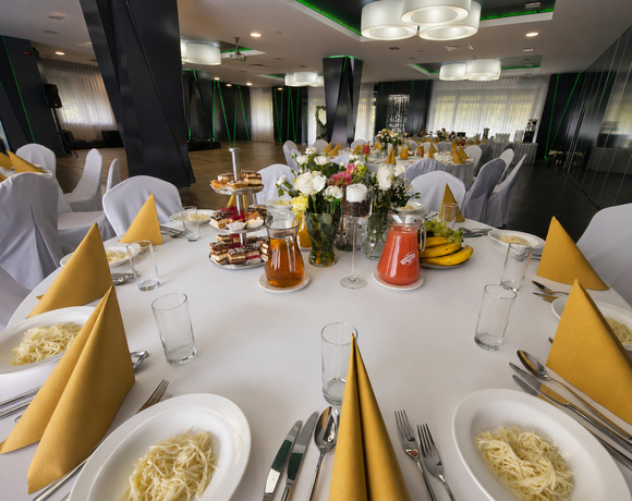 Sala bankietowa na przyjęcie: okrągły stół z białym obrusem, żółte serwety, makaron, bukiet kwiatów, ciasta i soki.