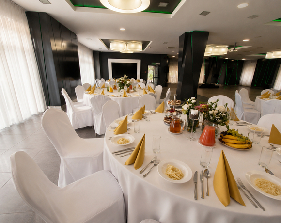 Elegancka sala bankietowa z okrągłymi stołami w białych obrusach, złotymi serwetkami, pokrytymi krzesłami, bukietami, dzbankami i półmiskiem owoców.