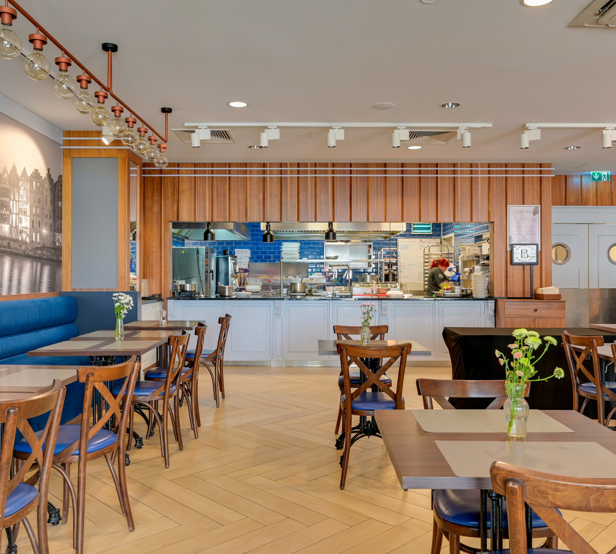 Bright hotel restaurant with open kitchen and chef at pass, blue banquette, wooden tables, globe pendant lights and fresh flowers