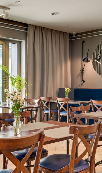 Heller Frühstücksraum im Hotel mit Holztischen und Stühlen, blauem Samtsofa, rundem Spiegel, Pflanzen und Blumenvasen.