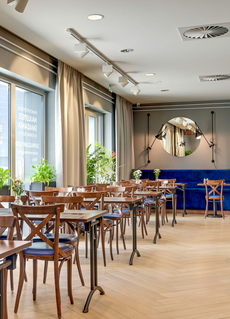 Jasna sala restauracyjna w hotelu z drewnianymi stolikami, niebieską kanapą, dużymi oknami, roślinami i okrągłym lustrem.