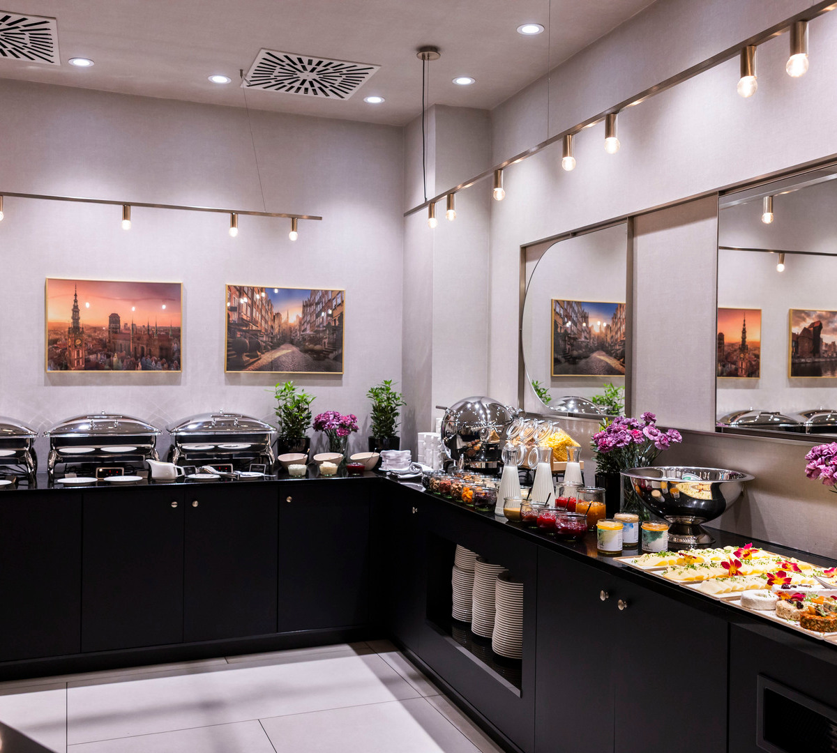 Elegantes Hotelbuffet mit Warmhaltern, Käse‑ und Salatplatten, Kaffeeautomaten, frischen Blumen und Stadtmotiven