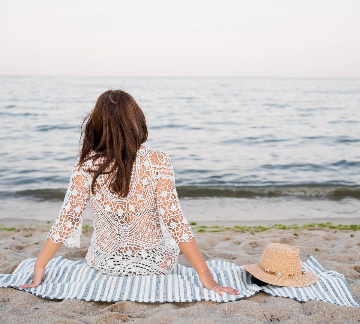 Kobieta w białym szydełkowym narzucie siedzi na pasiastym ręczniku plażowym, patrząc na spokojne morze; słomkowy kapelusz leży obok na piasku.