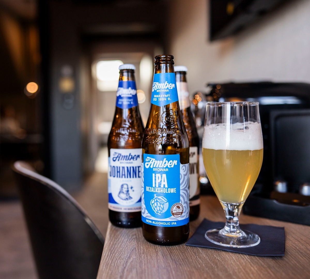 Alkoholfri IPA-flaskor med blå etiketter och ett grumligt, skummande öl i glas på ett träbord i en mysig bar