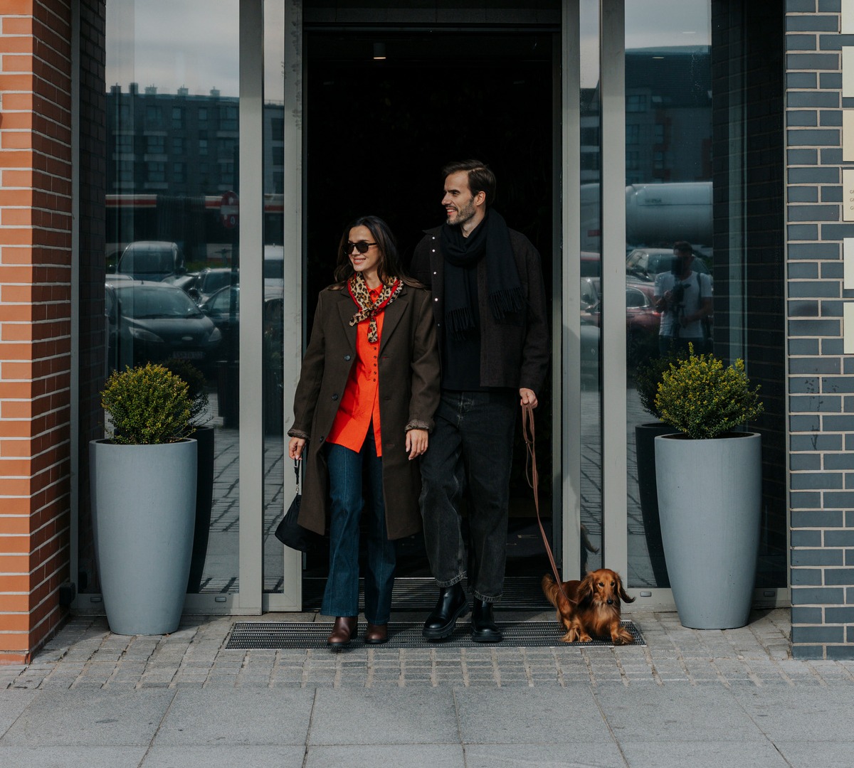 Par med liten hund går ut av firestjerners Grano Hotel gjennom glassdører foran moderne murfasade og store plantekasser.