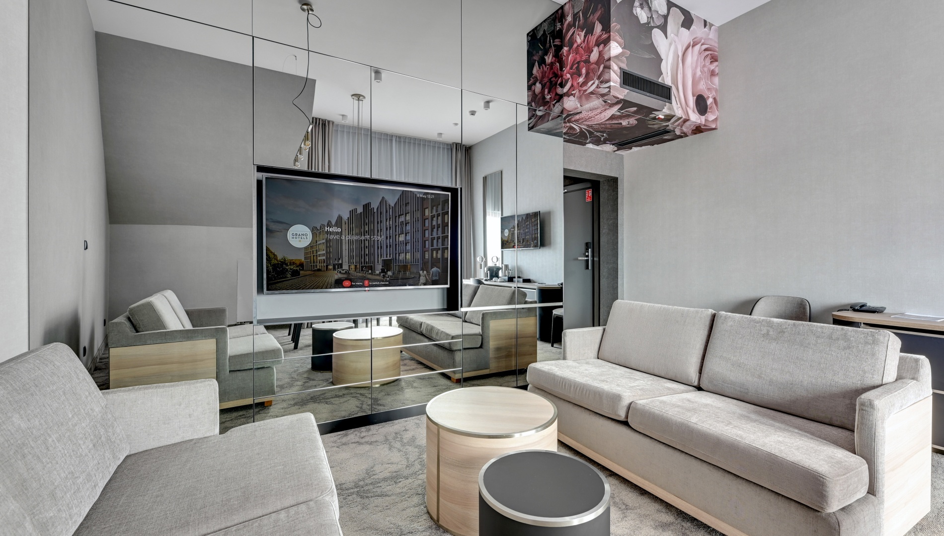 Moderne suite-lounge med grå sofaer, speilvegg med TV, runde tre- og svarte bord, blomsterdekor i taket, skrivebord og gulv-til-tak-gardiner