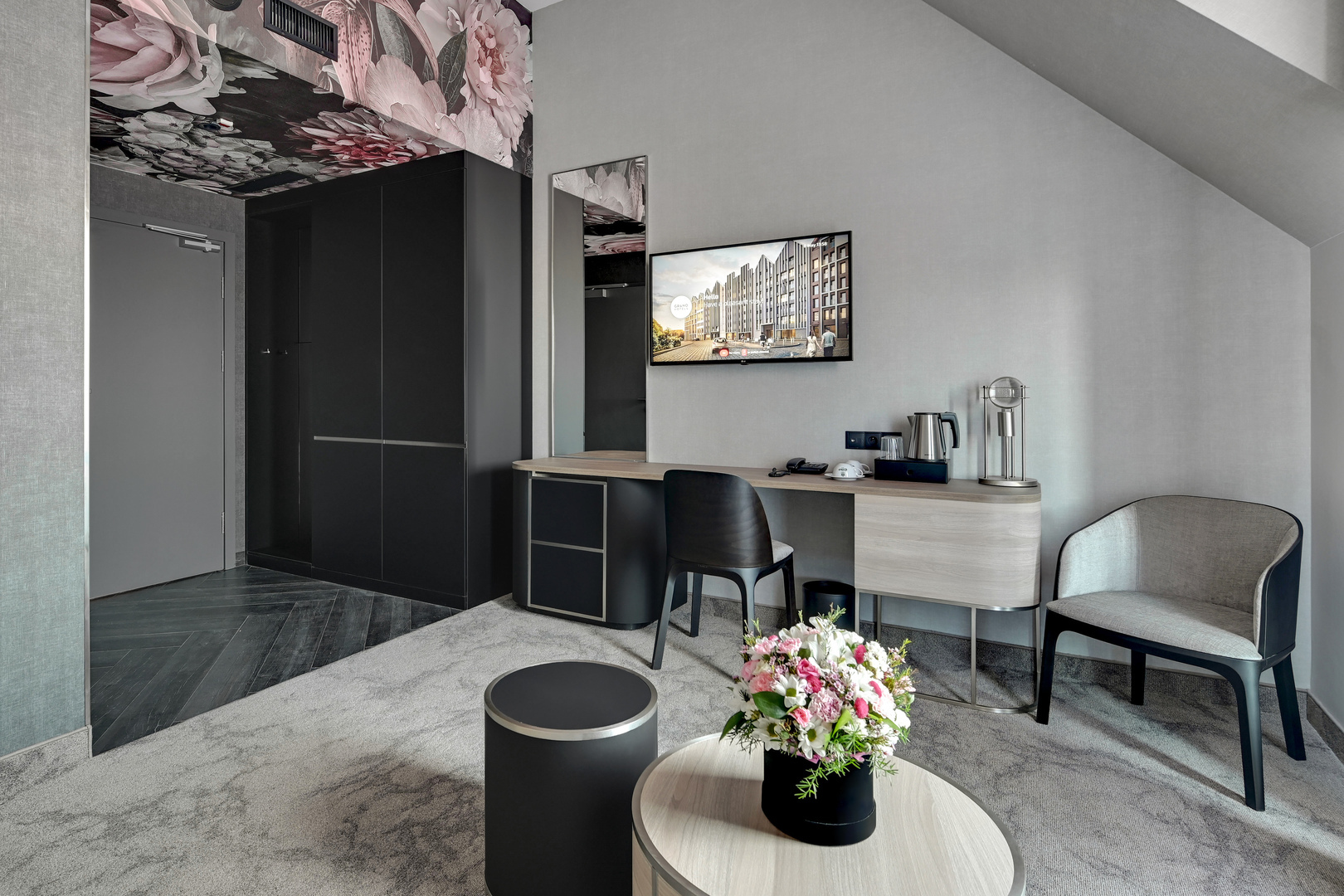 Moderne hotell-sitteområde med skrivebord, vegg-TV, vannkoker, lenestol, blomsterdekor i taket og bukett.