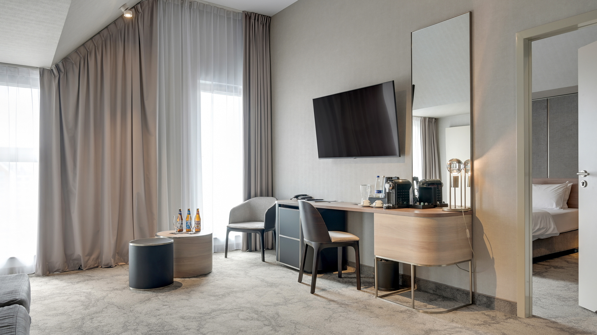 Nowoczesny pokój hotelowy z telewizorem na ścianie, biurkiem z ekspresem do kawy, miejscem do siedzenia, dużymi zasłonami i widoczną sypialnią