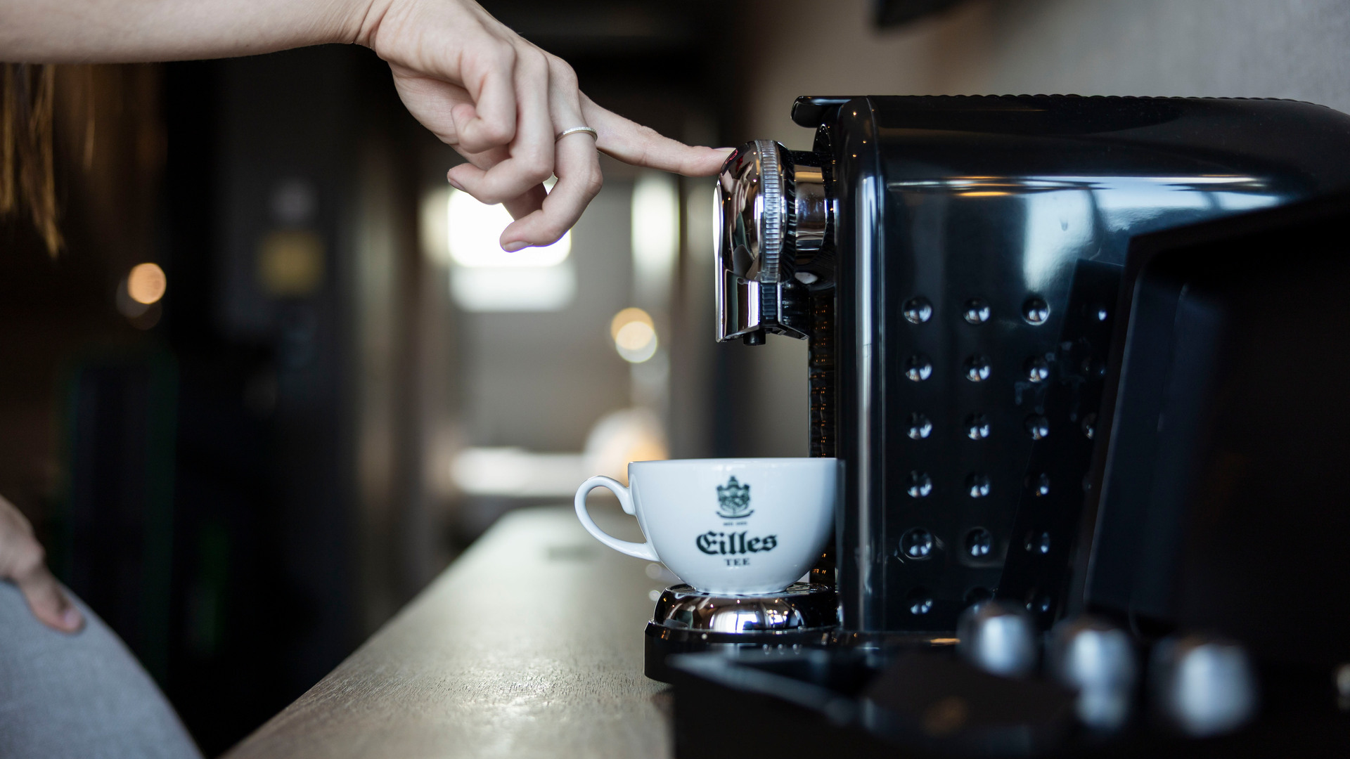 Hand trycker knapp på svart espressomaskin över en vit kopp på hotellens kaffestation