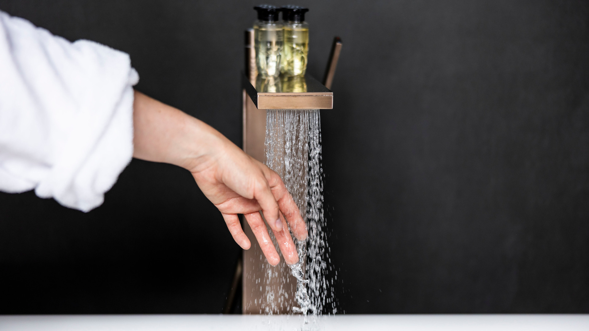 Hand under modern vattenfallsbadkarskran, hotelltoalettartiklar på kranen, synlig badrockärm