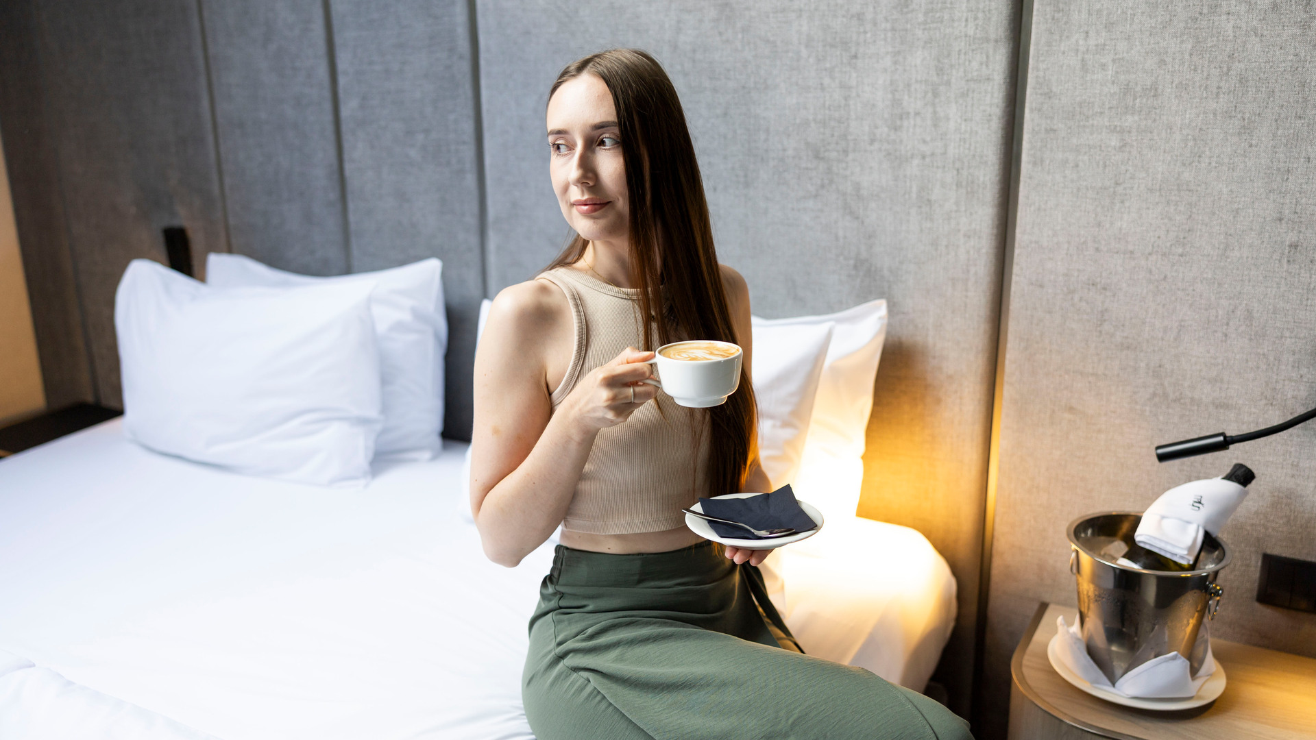 Kvinna på hotellsäng med latte och fat, grått stoppat sänggavel, varm sänglampa och champagnehink på sängbord