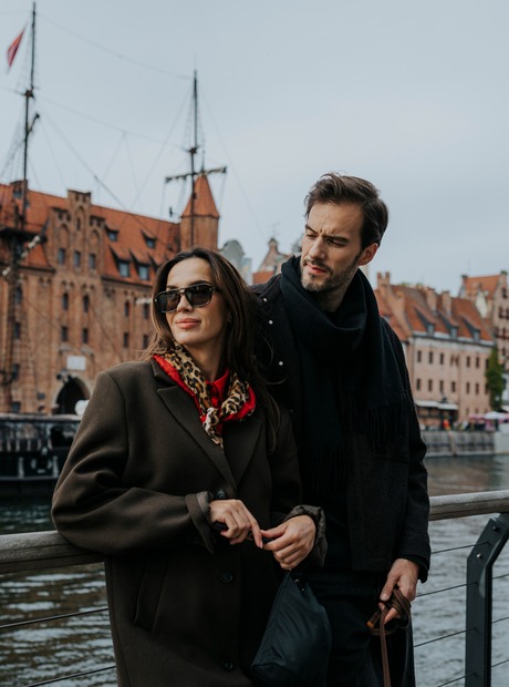 Par som går langs bryggen foran historiske rødmurte hus, gammel havnekran og et fortøyd seilskip
