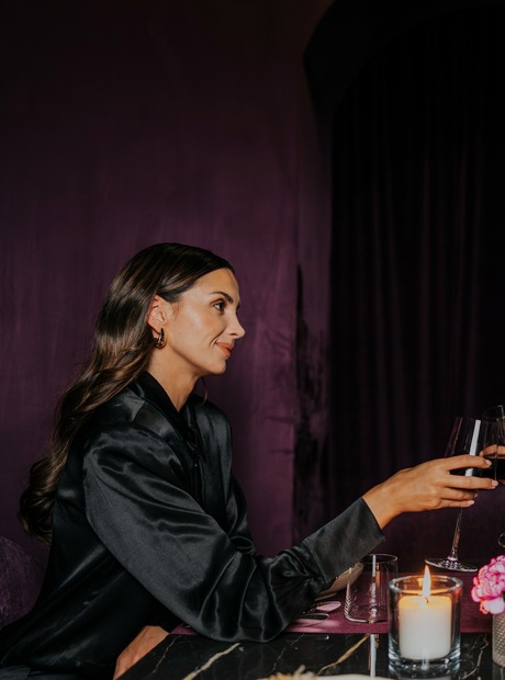 Paar in eleganter Kleidung stößt mit Rotwein an einem kerzenbeleuchteten Tisch mit Blumen und Vorspeise, violetter Samtvorhang, intime Restaurantatmosphäre