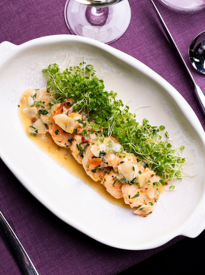 Gebratene Garnelen in Knoblauch‑Kräutersauce mit Mandelblättchen und frischem Mikrogrün, serviert neben einem Weinglas