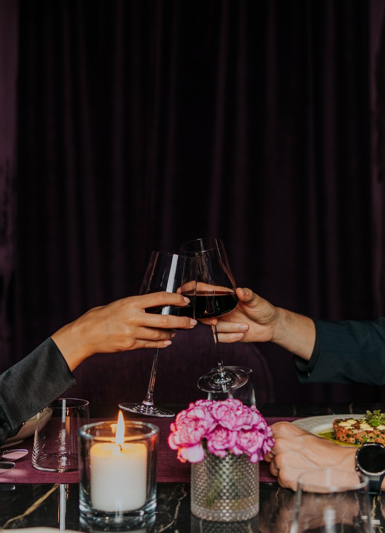 Par i elegant antrekk skåler med rødvin ved et stearinlysbelyst bord med blomster og forrett, lilla fløyelsbakgrunn, intim restaurantstemning