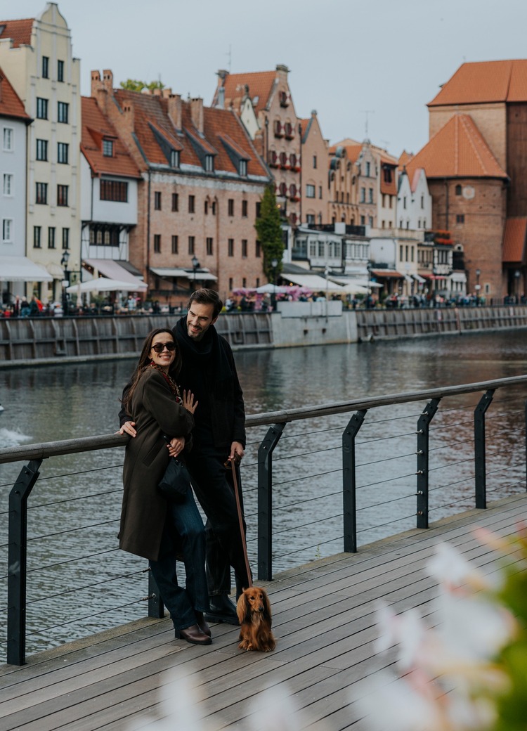 Par går langs havnepromenaden foran historiske mursteinsbygg, med kafeer, båtturer og en liten hund ved føttene