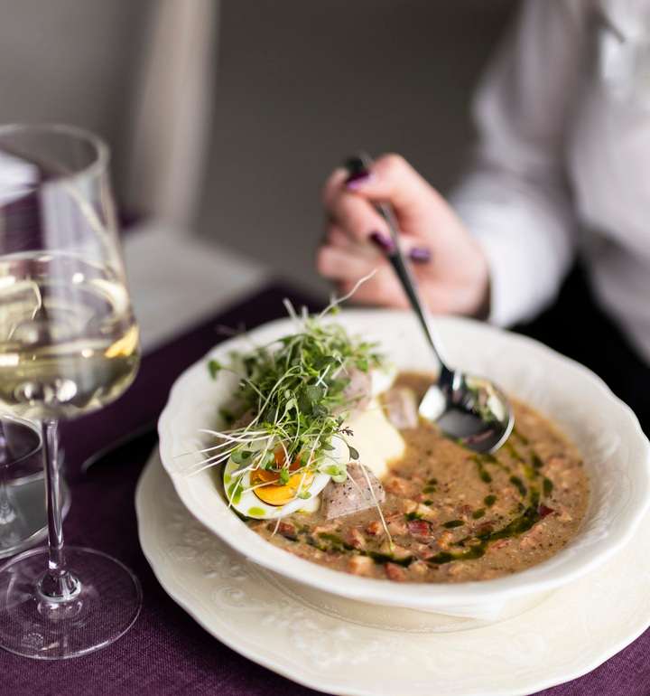 Cremige Suppe mit halbiertem Ei, Sprossen und Kräuteröl in weißer Schale, dazu Wein und eine Hand mit Löffel.