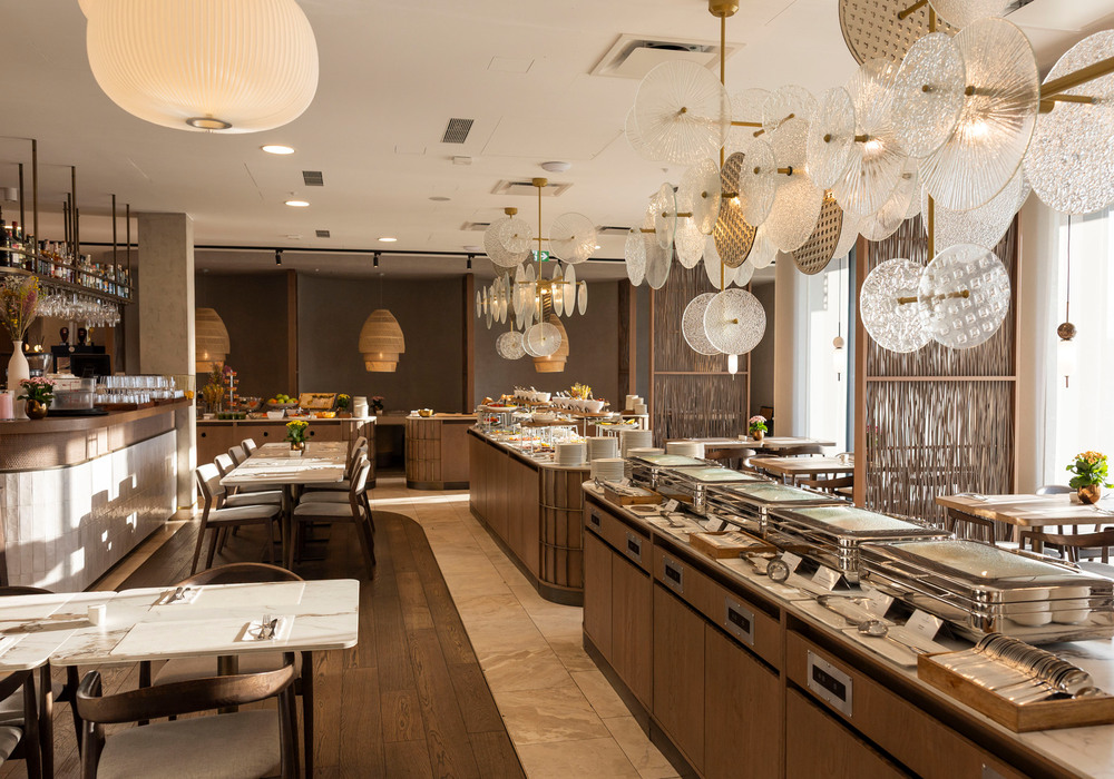 Elegancka restauracja hotelowa z wyspą bufetową, marmurowymi stolikami, barem, ciepłym oświetleniem i dekoracyjnymi szklanymi żyrandolami.