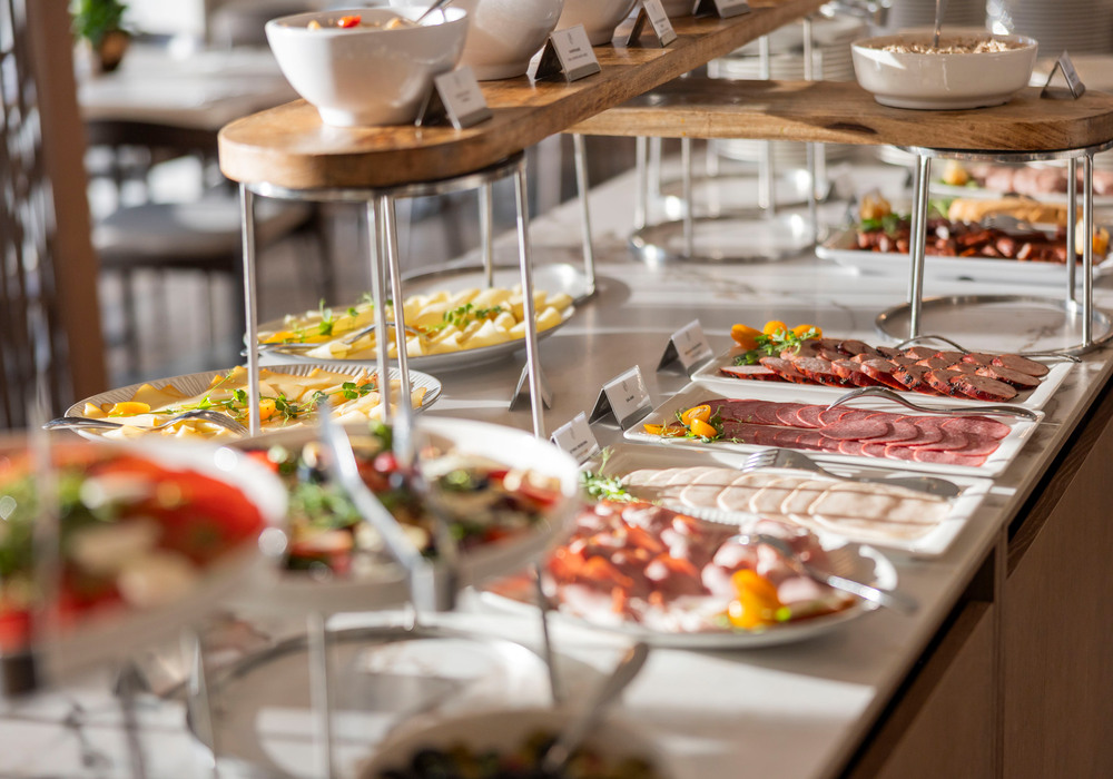 Bufet śniadaniowy w hotelu z drewnianą półką, talerzami wędlin, serów, sałatek i misami, jasne, eleganckie ustawienie