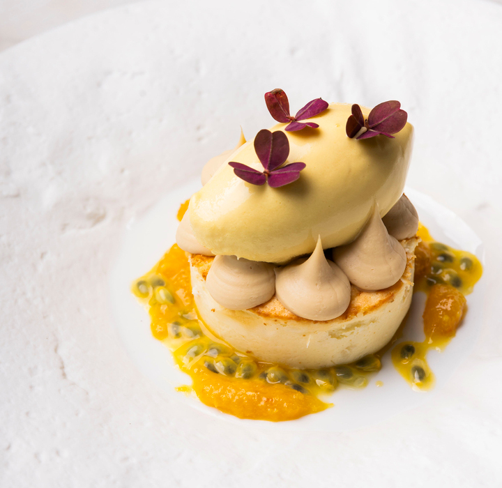 Tropikalny deser na talerzu: okrągły biszkopt, beżowe kremowe kopułki, purée z mango, nasiona marakui i żółty sorbet z fioletowymi mikroliśćmi.
