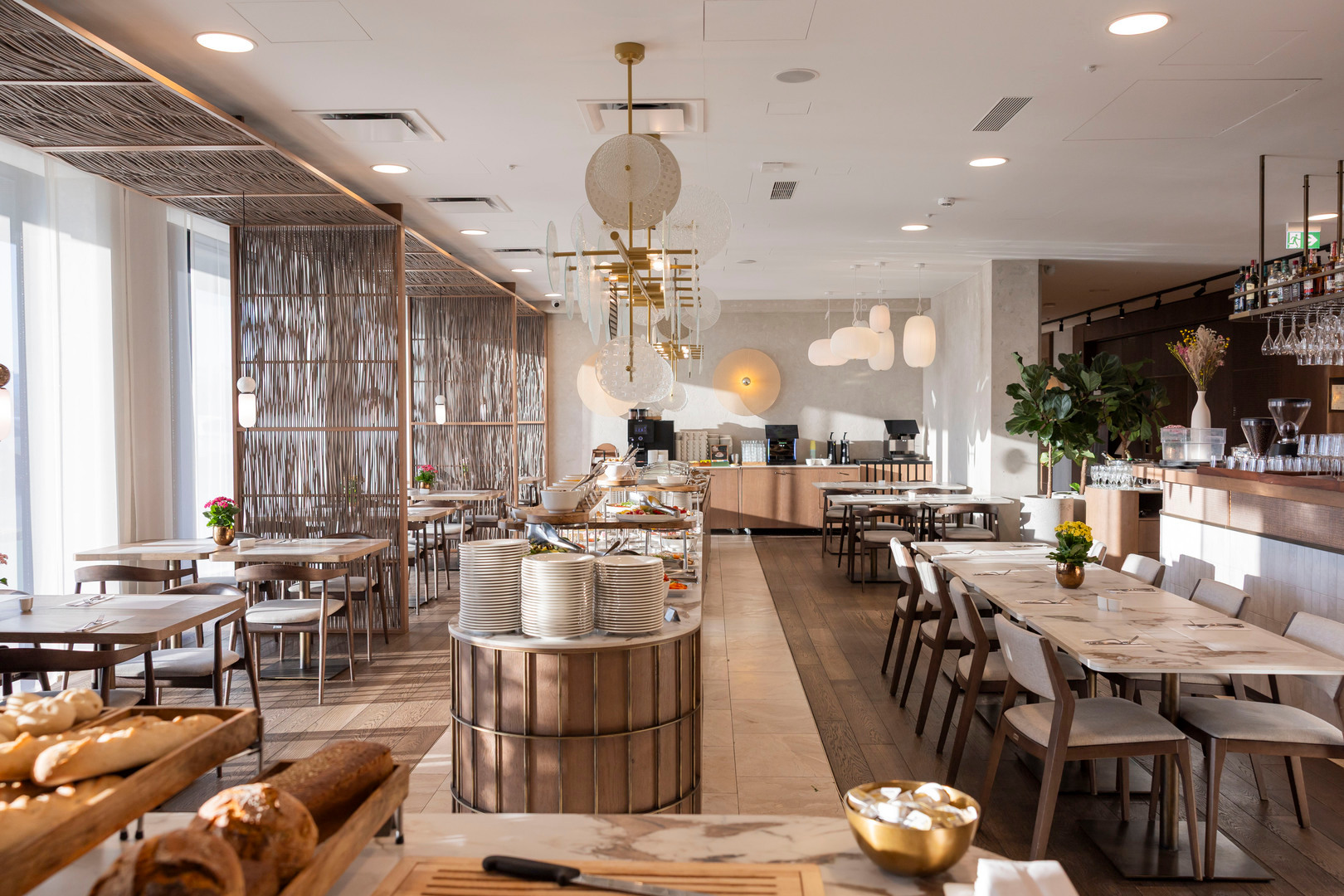 Helle, moderne Hotelfrühstückslounge mit Buffet, frischen Backwaren, gestapelten Tellern, Holztischen, Pendelleuchten und Bar