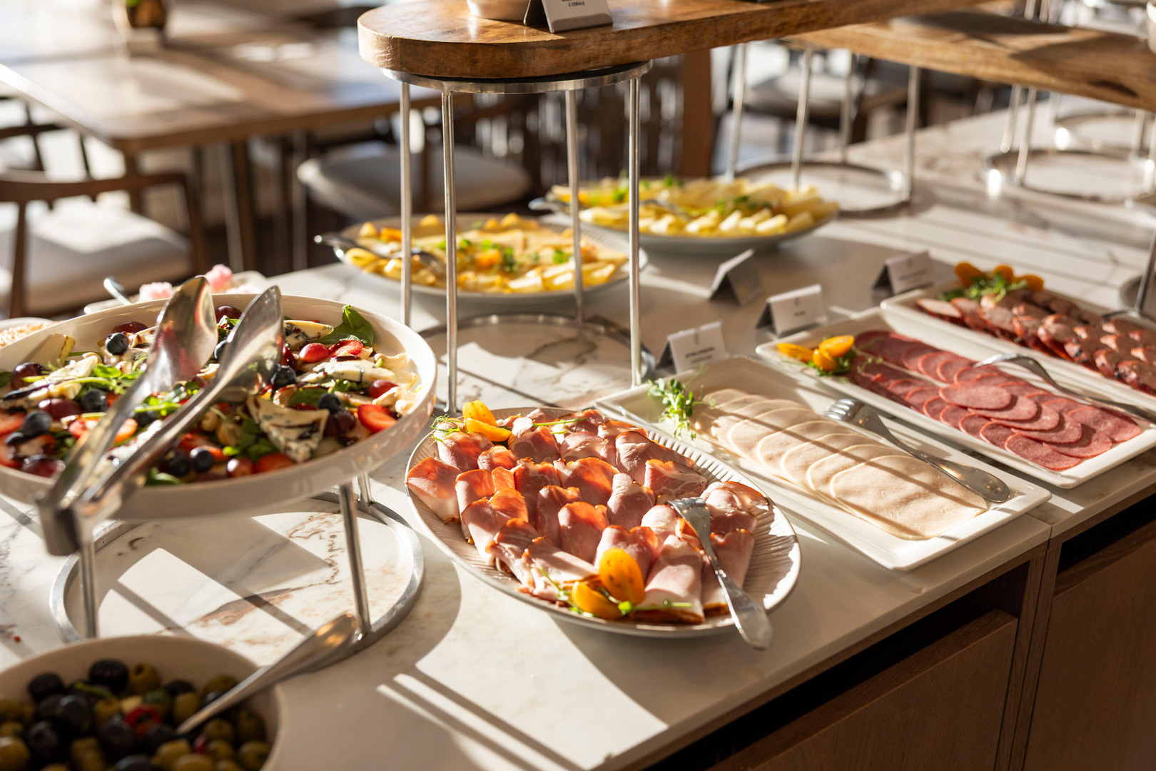 Frühstücksbuffet mit angerichteten Aufschnittplatten, Käse, fruchtigem Salat und Oliven auf sonnigem Marmor