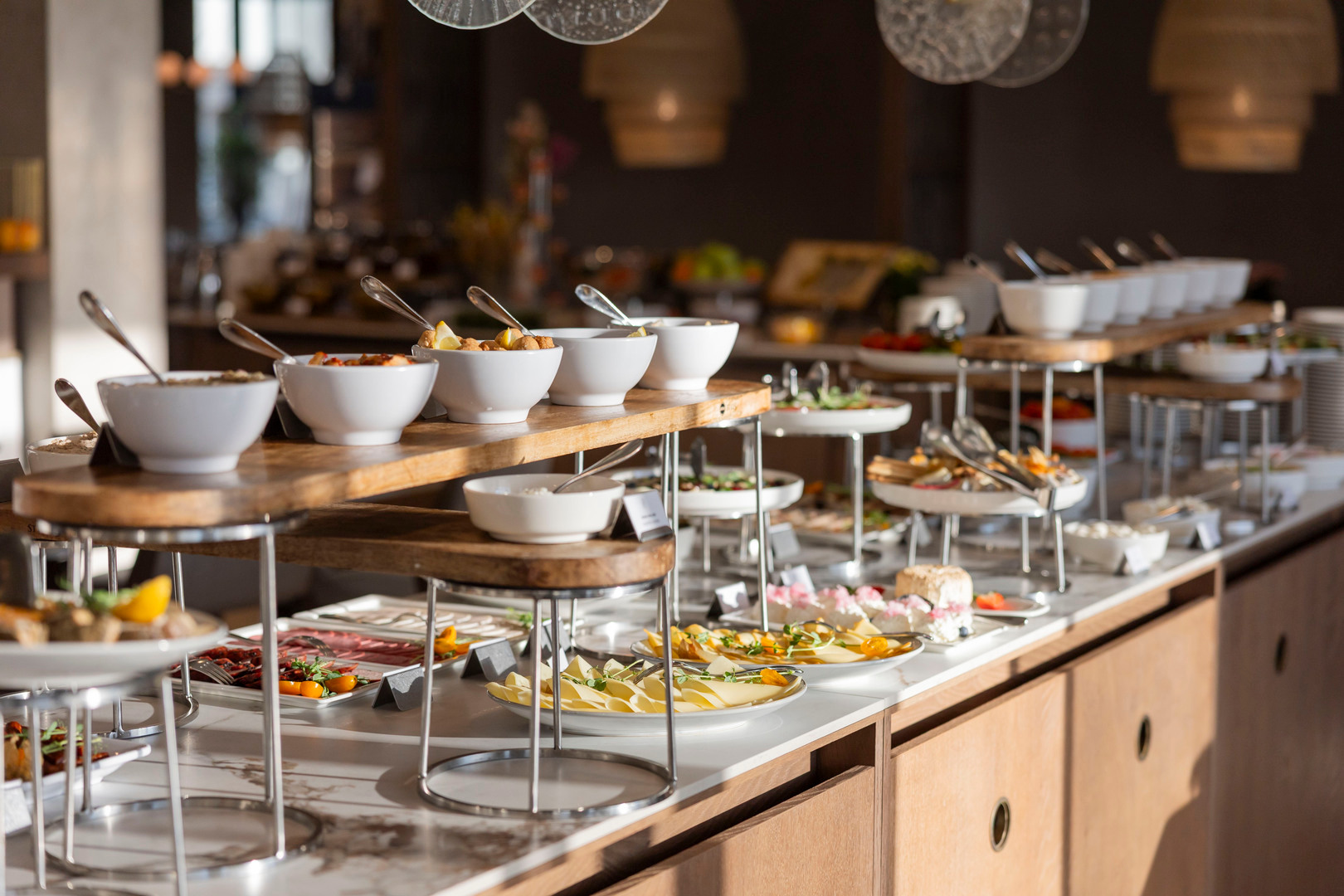 Hotel-Frühstücksbuffet mit Käse, Aufschnitt, Salaten und Beilagen in weißen Schalen auf Holzständern