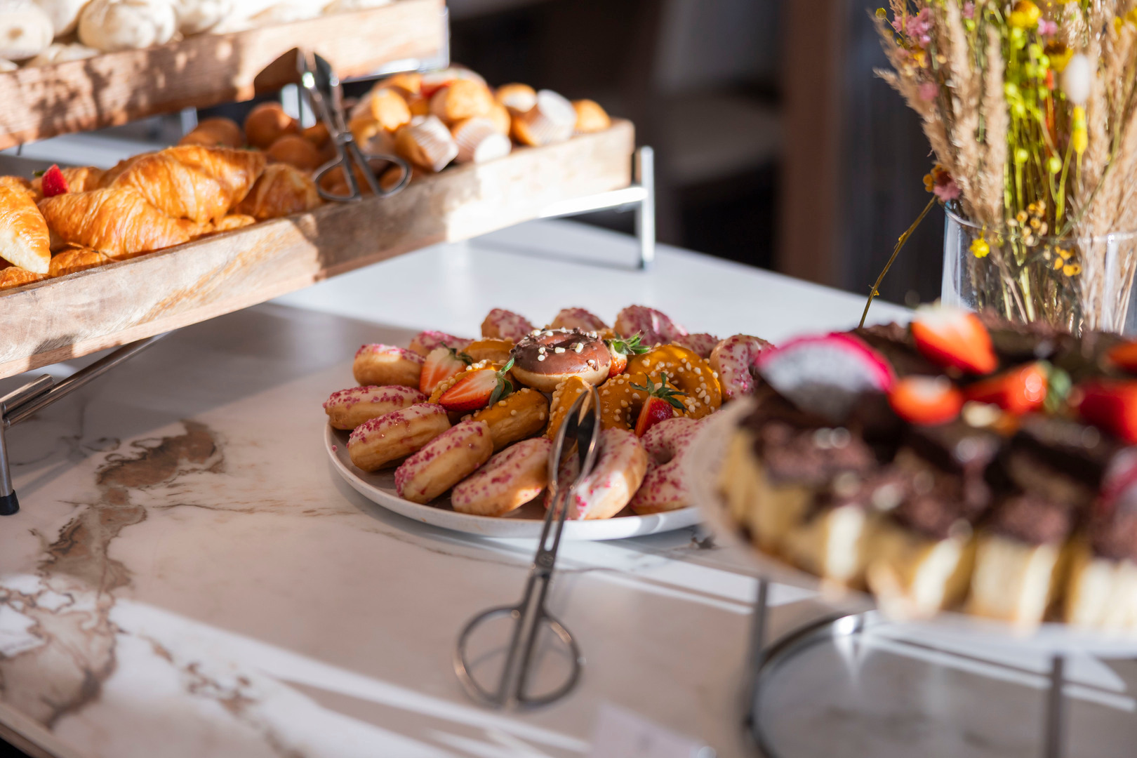 Frühstücksbuffet mit goldenen Croissants, bunt bestreuten Donuts und frischen Erdbeeren auf Marmorplatte.