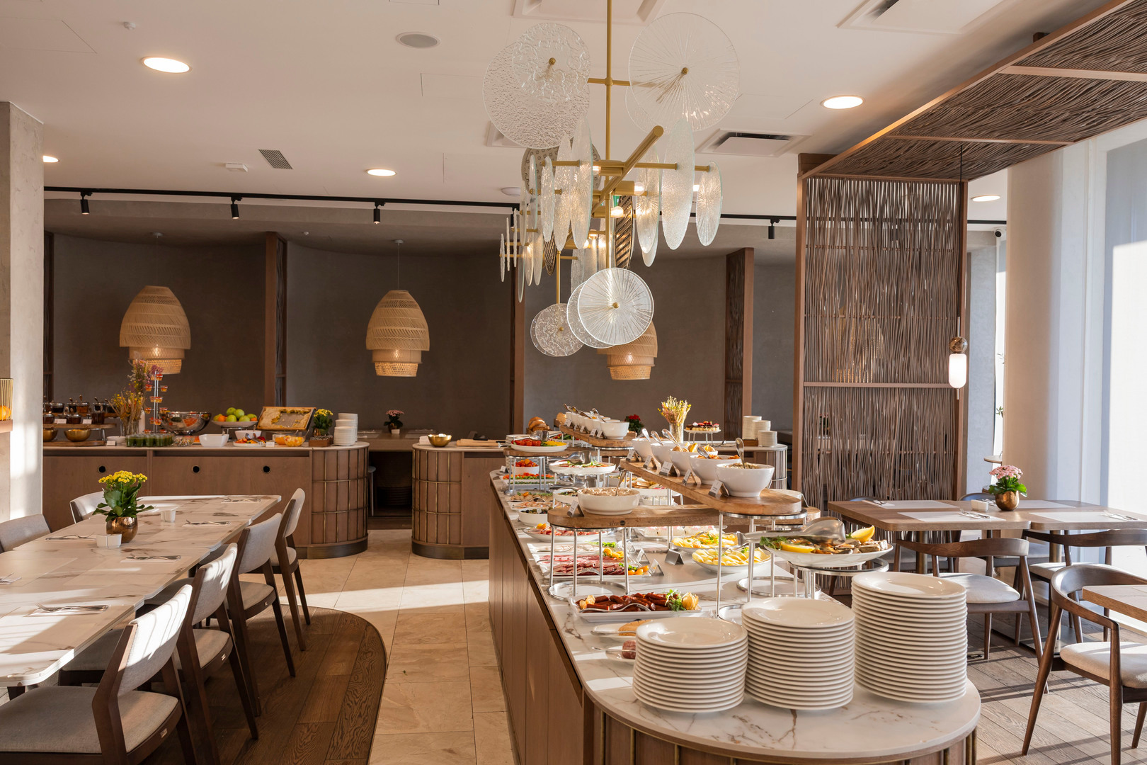 Elegantes Hotel-Buffet auf einer Marmorinsel mit gestapelten Tellern, frischem Obst und Aufschnitt, warmen Holzmöbeln und geflochtenen Hängeleuchten