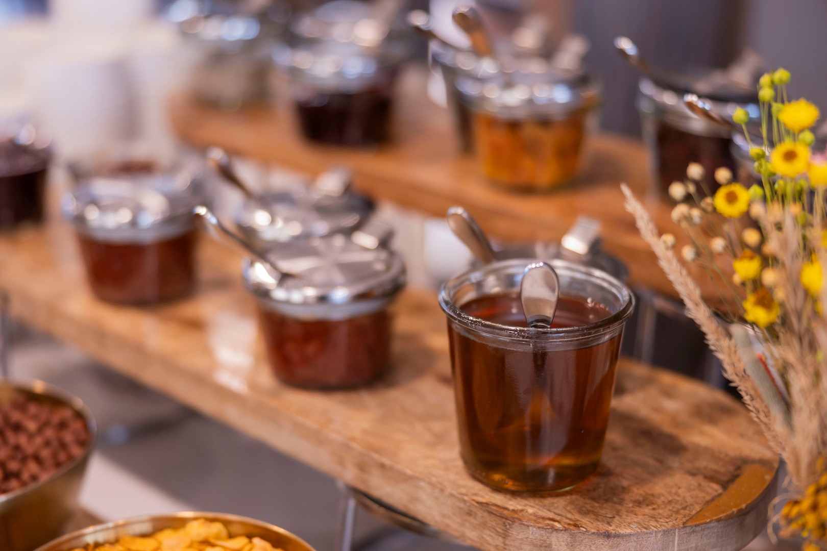 Frühstücksbuffet mit Gläsern Honig und Fruchtaufstrichen, Löffel auf rustikalem Holzbrett, gelbe Blumen