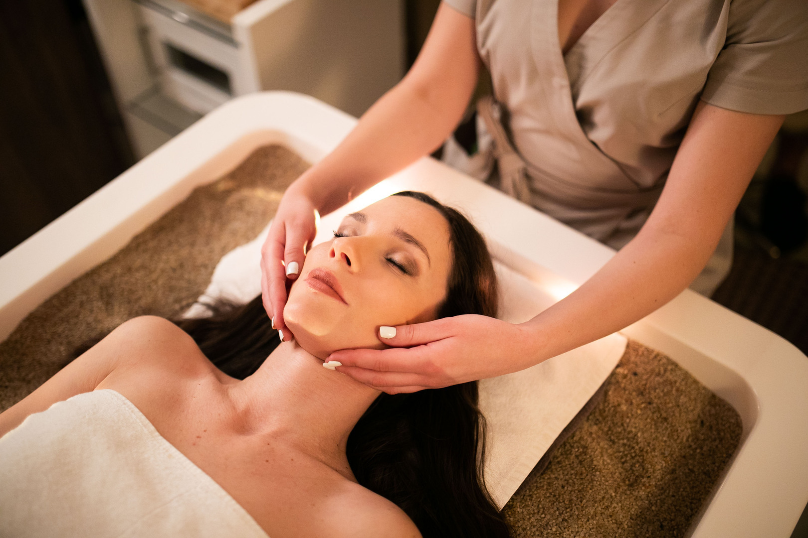 Guest receives a relaxing facial massage on a spa bed, therapist's hands gently cradle her jaw in warm light
