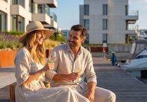 Par nyter hvitvin på bryggen ved moderne sjønære leiligheter og en fortøyd yacht, solfylt og avslappet stemning.