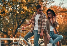 Couple with a bicycle in a sunlit autumn park, stylish casual attire and a relaxed, romantic vibe for guests.