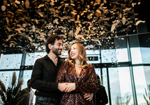 Couple in evening attire embracing under confetti in a stylish hotel lounge with floor-to-ceiling windows