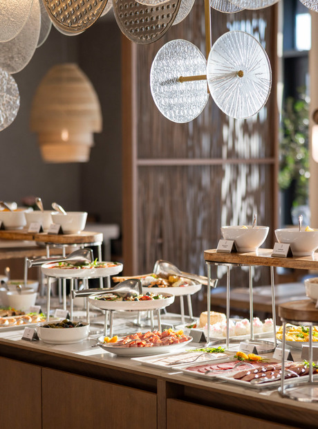 Hotelbuffet mit Salaten, Aufschnitt, frischem Obst und Schalen auf Holzpodesten unter dekorativen Hängeleuchten.