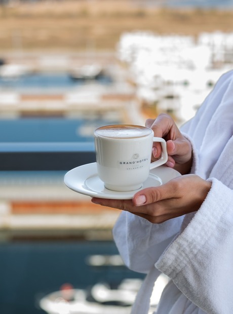 Gast im weißen Bademantel hält Hotelkaffee auf dem Balkon mit Blick auf Marina und Yachten, entspannter Spa-Morgen