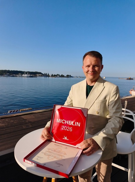 Mann im beigefarbenen Anzug hält die Michelin-Auszeichnung 2026 auf einer Terrasse am Wasser mit ruhigem Meer und klarem Himmel