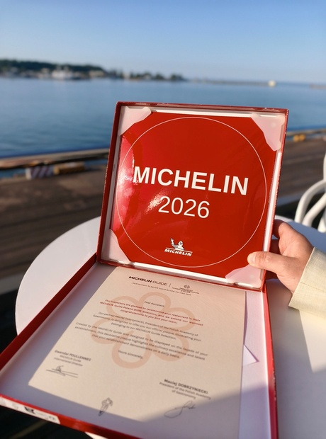 Czerwona tablica MICHELIN 2026 w otwartym pudełku z certyfikatem, dłoń trzymająca ją na nadmorskim tarasie.