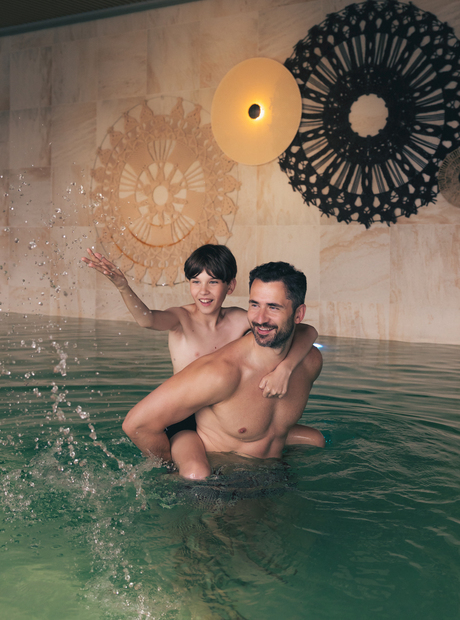 Vater trägt Sohn auf dem Rücken im hoteleigenen Hallenbad, planscht im Wasser unter warmer Wandbeleuchtung und Dekor