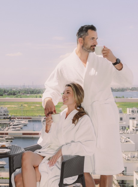 Para w białych szlafrokach na hotelowym balkonie pije kawę, z ciastkami na stoliku, w tle marina i nowoczesne apartamenty, relaksujący poranek