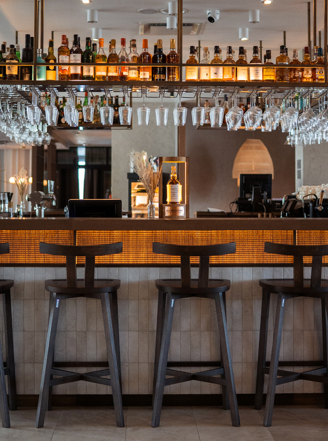 Stylowy bar hotelowy z podświetlanym plecionym frontem, wysokimi stołkami, wiszącymi kieliszkami i półką z alkoholem
