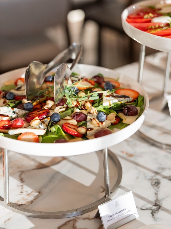 Buffet-Salat auf weißer Servierplatte mit Blattsalat, Erdbeeren, Blaubeeren, Nüssen und weichem Käse, Servierzange auf Marmor